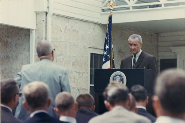 President Lyndon Johnson stands at a podium to field a reporter's question at a press conference, probably at the Texas White House.