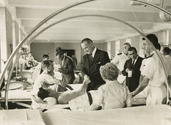 On a surprise visit to Bethesda Naval Hospital, President Lyndon Johnson shakes hands with John R. Kington, a Marine wounded in Vietnam. In the next bed is Donald Grove. Taking notes on this visit is White House correspondent Merriman Smith.