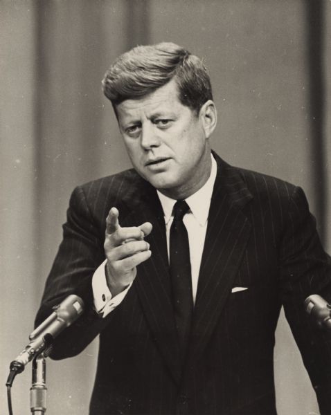 Waist-up portrait of President John F. Kennedy standing at a podium at his first press conference.