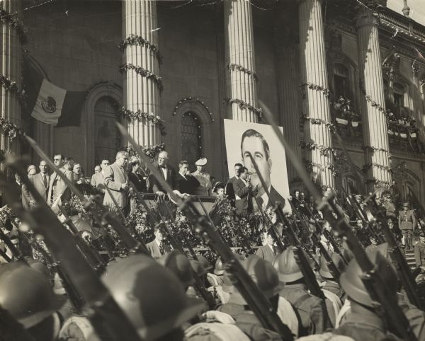 President Franklin Roosevelt and Mrs. Roosevelt on the officials platform during a visit to Monterrey, Mexico. Standing between them (and also pictured on the large banner) is Mexican president Avila.