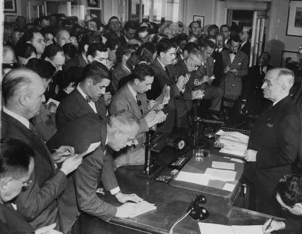 President Harry Truman meets a packed room full of reporters. Merriman Smith, the "dean of the White House correspondents and the donor of this image," can be seen above the reporter who is leaning on the President's desk.
