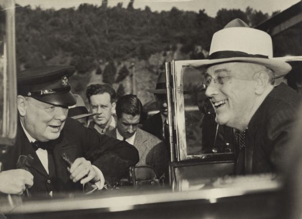 Prime Minister Winston Churchill greets President Franklin Roosevelt as he is about to exit his vehicle for their meeting at Quebec. This was their second meeting at Quebec at which decisions were made about the occupation of a defeated Germany, British support for the war in the Pacific, and continued aid to Britain. UPI reporter Merriman Smith, who is looking down at his notebook, can be seen between the two leaders.