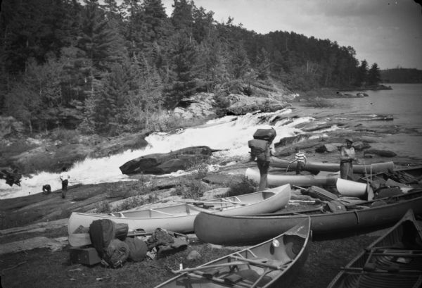 View of men and young boys with canoes pulled up on rocks near a rapids during an unidentified wilderness canoe trip led by Sigurd Olson.