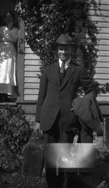 Sigurd Olson, posing proudly, as he leaves his Ashland home for study at the University of Wisconsin. A woman is standing in the doorway of a house behind him.
