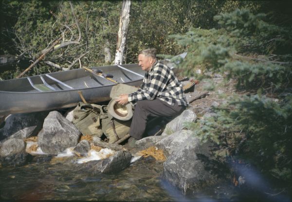 Supreme Court Justice William O. Douglas during a canoe trip with conservationist Sigurd F. Olson. He is sitting with his hat in hand beside a canoe pulled up on the shoreline.