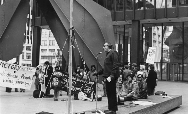 "Irwin Silber, an editor of the 'Guardian'," a radical weekly newspaper, speaking to an anti-war rally in a square near a large sculpture.