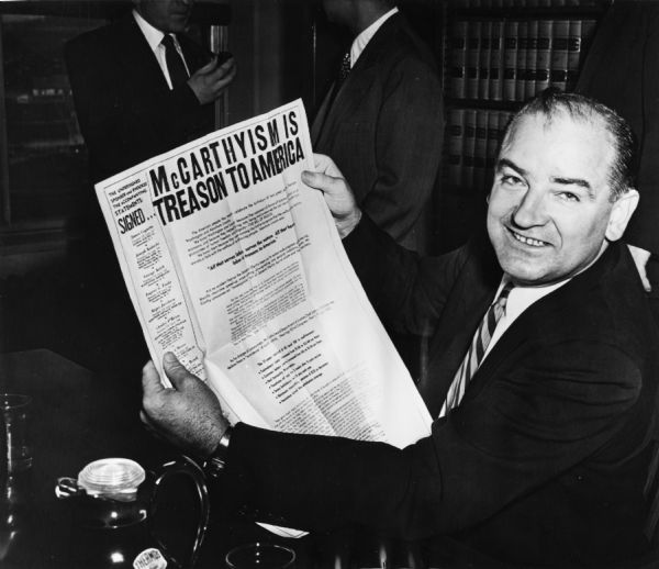 Senator Joseph R. McCarthy, laughing for the camera, displays a newspaper advertisement that proclaims: "McCarthyism is Treason to America."