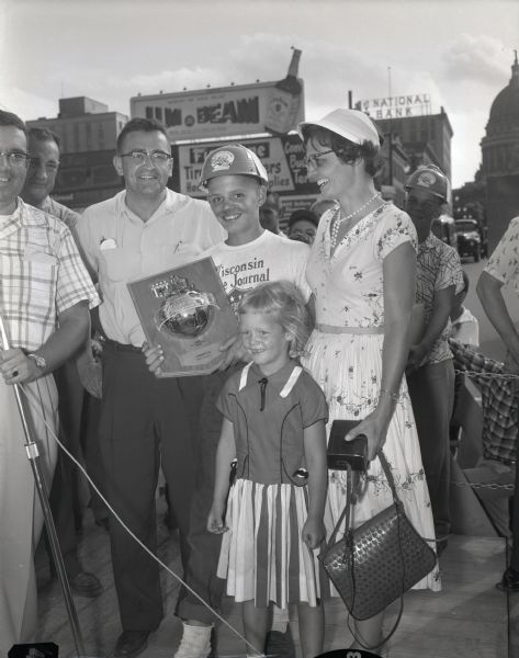 Madison Soap Box Derby winner Alan Strauss holding his trophy and standing with his family on East Washington Avenue. The Wisconsin State Capitol is in the background.