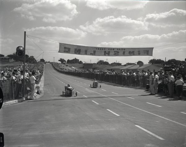 Finish line of the Soap Box Derby on Midvale Boulevard. Two cars are about to cross the line with crowds of people watching from behind fences along the sidewalks.
