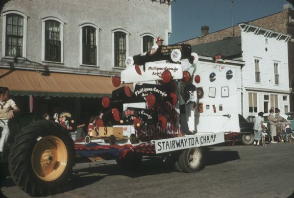 "Stairway to a Champ" float in a parade. The float includes the five cars raced by Van Steiner of Argyle along with his helmets and trophies.<p>Van won the Madison Soap Box Derby in the car at the top, marked "Wisconsin State Journal," and competed in it at Derby Downs, Akron, Ohio. Van stands at the back wearing his champion shirt from Akron. His brother sits in the top car.</p>