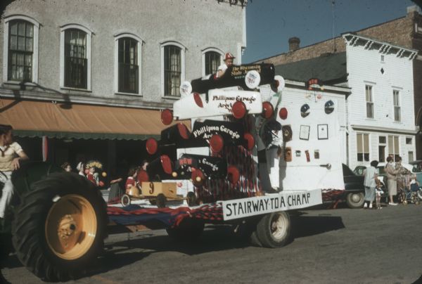 "Stairway to a Champ" float in a parade. The float includes the five cars raced by Van Steiner of Argyle along with his helmets and trophies.<p>Van won the Madison Soap Box Derby in the car at the top, marked "Wisconsin State Journal," and competed in it at Derby Downs, Akron, Ohio. Van stands at the back wearing his champion shirt from Akron. His brother sits in the top car.</p>

