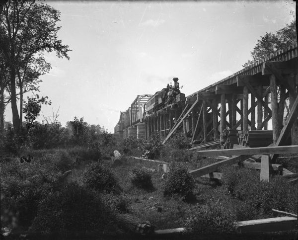 View from below of wood burning locomotive and single car on elevated railroad bridge and approach trestle, somewhere along the upper Mississippi River. Large timbers are stacked and strewn below the tracks, and a number of men are posing alongside the train.