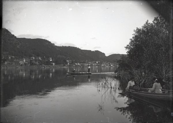 Oblique view from the Mississippi River. Two young women are in a rowboat near the shoreline, and two young men are fishing in the river. Alma at the foot of large bluffs can be seen on the opposite shoreline.