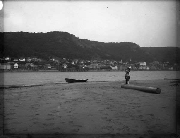 View of Alma across river from sandbar. A rowboat is on the sandbar, and on the right a father and child are standing posed each with one foot on a log.