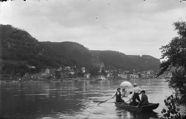 Four men and one woman in a rowboat on the Mississippi River. In the background is the town of Alma at the foot of bluffs. The woman is holding an umbrella.