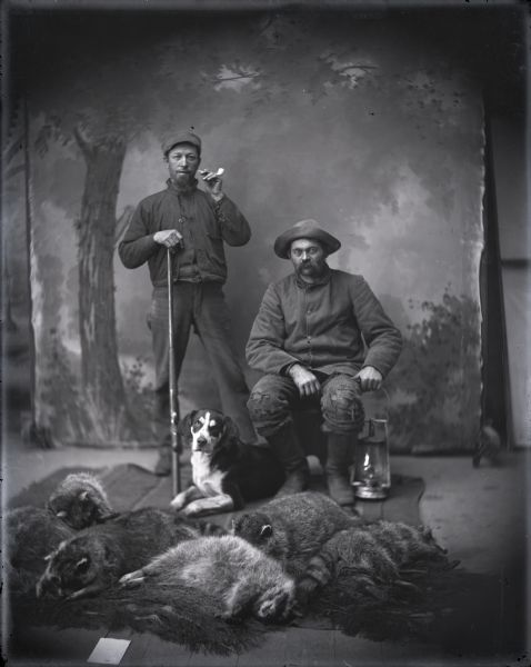 Studio portrait in front of a painted backdrop of two hunters, Jacob Gross, sitting and holding a lantern, and Henry Herold, standing and holding a rifle and pipe. A hound dog sits near them, and in the foreground a group of dead raccoons are displayed. The portrait was taken the morning after a successful hunt. Gesell staged the scene to look like a night in the woods.