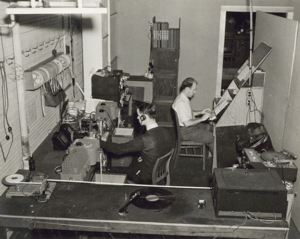 Elevated view of two men operating electronic equipment at the U.S. Naval Training School (Radio). One of the men is wearing headphones, and there is a long-playing (LP) record on a turntable in the foreground.