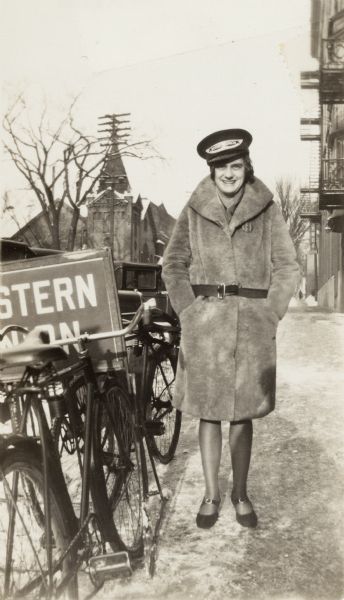 A woman stands outside of the Western Union office at 650 State Street. She is next to a Western Union sign and several bicycles, and there is snow on the ground. She is wearing one of the messenger boy's caps. On the right is the YWCA. In the left background are Calvary Lutheran (713 State Street, on the corner of Lake and State Streets) and Saint Paul's Catholic Church (717 State Street).