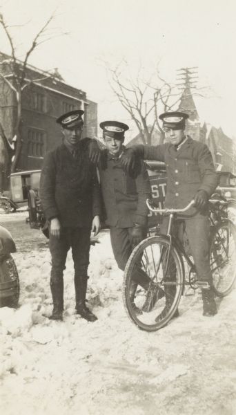 Three Western Union messenger boys pose with a bicycle in front of the Western Union sign at 650 State Street. There is snow on the ground. The messenger boys were usually university students.