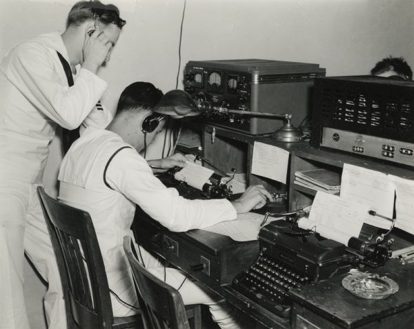 One Naval student operates a telegraph while another student observes, listening with earphones, at the U.S. Naval Training School (Radio). A typewriter is in the right foreground.