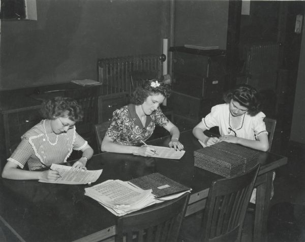 Three women sitting at a table appear to be proofreading documents at the U.S. Naval Training School (Radio).