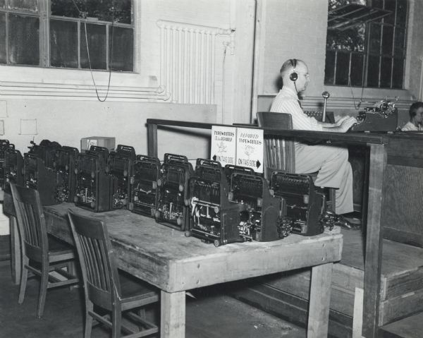 A table top is filled with typewriters at the U.S. Naval Training School (Radio). Two signs identify which need repair, and which have been repaired. In the background on a raised platform is a man sitting at a typewriter wearing headphones. A student appears to be sitting behind him.