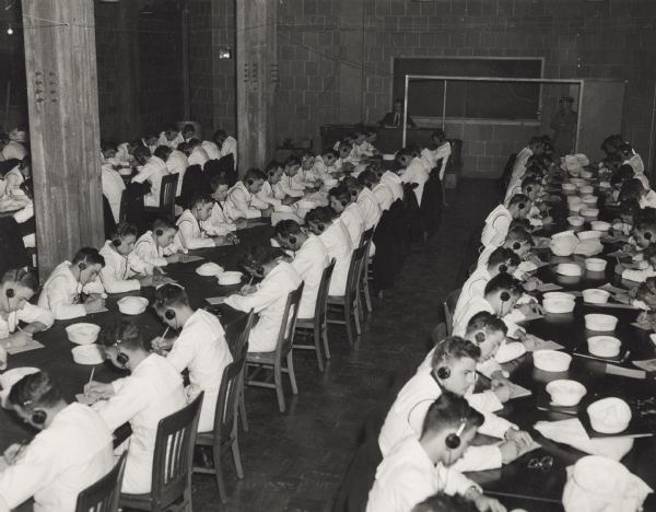 Naval students sit together at long tables and write messages while listening to earphones at the U.S. Naval Training School (Radio). In the background a man sits at a desk against the wall.