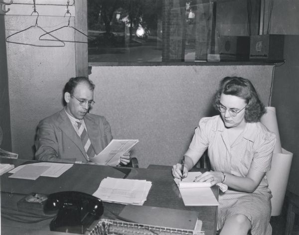 A man is reading at a desk, while a woman writes on a pad of paper seated nearby in an office at the U.S. Naval Training School (Radio).