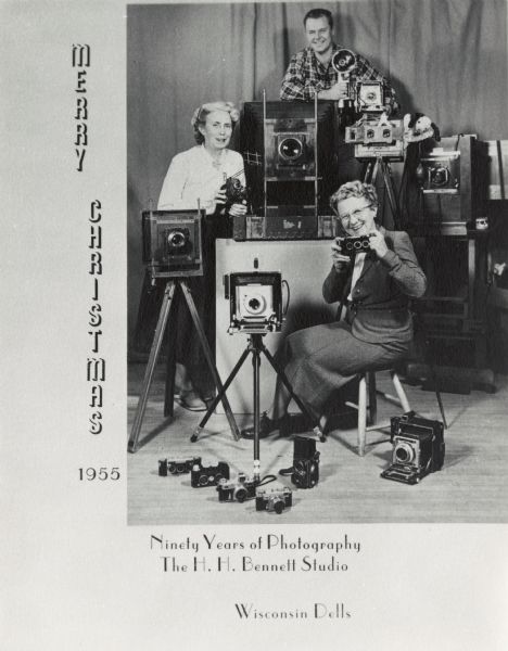 H.H. Bennett Studio Christmas portrait of two women, Miriam and Ruth, daughters of H.H. Bennett, and one man, Oliver Reese, posed with many cameras, some on tripods. To the left of the photograph is the vertical text "MERRY CHRISTMAS" and just below that is the year "1955." Below the photograph more text reads "Ninety Years of Photography, The H. H. Bennett Studio, Wisconsin Dells."