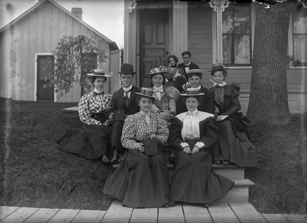 A group portrait of the Shutterbugs Camera Club. First row, left to right: Suzy Dempsey, Jennie Dempsey; Second row, middle, Anna Dempsey; porch step, Libby(?), Melendy and James Dempsey. They are posed on the steps to a house.