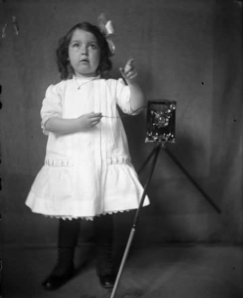 A young girl, Tannisse Taylor, poses with a camera on a tripod. She is wearing a white dress, black stockings, black shoes and has a white bow in her hair. She is holding the shutter release in her hand, and is probably the daughter of the photographer.