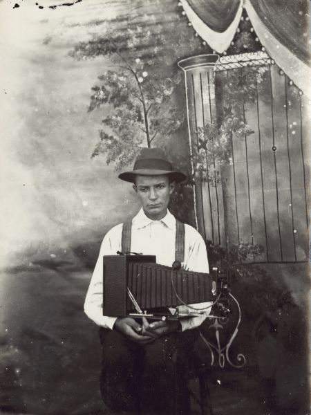 A studio portrait of a man posing with a camera in front of a painted background. He wears a white shirt, suspenders and a hat.
