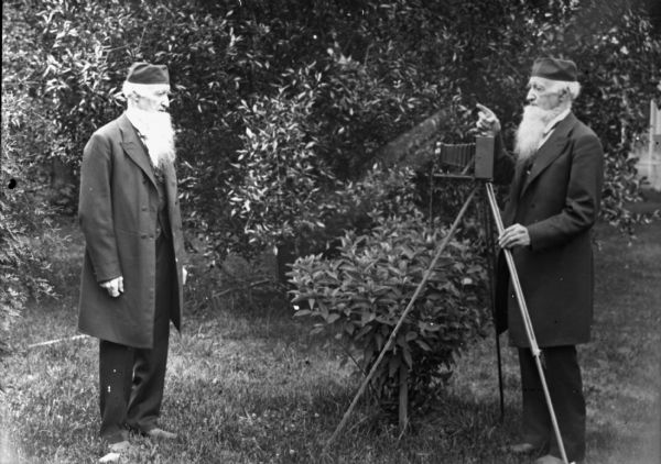 A photograph of a man, M. Newman, taking a picture of himself outdoors. He appears twice. The photographer used a double exposure to create the picture. He is wearing a dark suit, coat and cap, and has a long white beard.