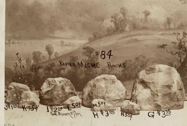 A photographic postcard of studio props (#84 PAPIER MACME ROCKS) in front of a painted backdrop. There are 8 different rocks in different sizes. The prices range from $3.75 for the largest rock, (G), to 50 cents for the 3 smallest rocks, (L, M and N).