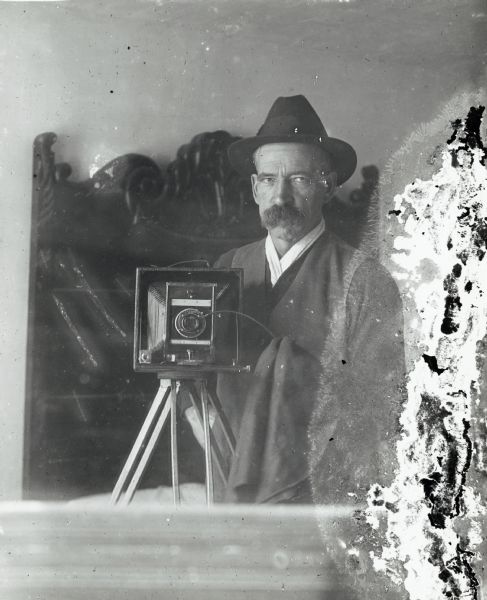 A photographic self-portrait of David Gander taken in a mirror. He is posed with a camera and is wearing a jacket and a hat. In the background appears to be a headboard of a bed, perhaps the mirror is on a dresser in a bedroom. David Gander was a photographer located in Gays Mills, Wisconsin.