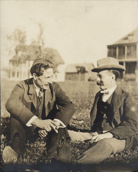 Two men sitting in the grass and visiting. Both men appear to be holding cigars. One man is wearing a jacket, vest, bowtie and hat. The other man is wearing a jacket, vest and tie. Homes appear in the background. The text on the back of the photograph reads: "Will Henry McFetridge and Louis Claude, family friend and Baraboo, Wisconsin architect. The Claude family lived on the shores of Devil's Lake and evidently played a role in Will Henry championing Devil's Lake as a State Park." Devil's Lake became a State Park in 1911.