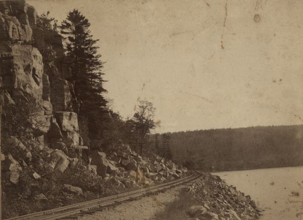 A view of the shoreline of Devil's Lake with railroad tracks in the foreground. On the back of the photograph is the photographer's information: "Sim Mould, Photographer, Views of Lake and Dells, Baraboo, Wis."