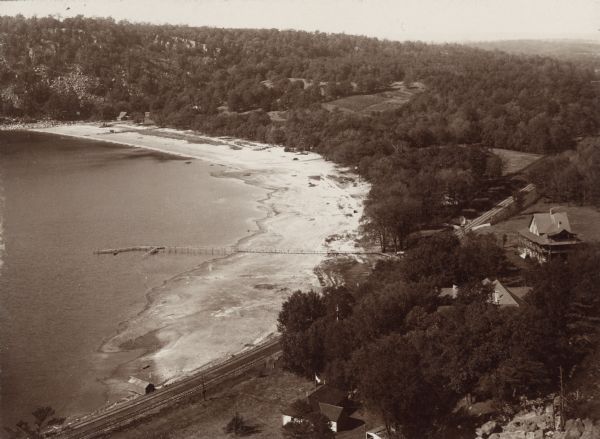 Elevated view of the north end of Devil's Lake and Cliff House. A number of buildings are around the shoreline with railroad tracks in the foreground. A long pier crosses the beach into the lake.
