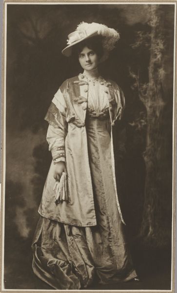 Full-length studio portrait of Blanche Budd McFetridge in front of a painted backdrop. She is wearing a dress with a matching jacket and a hat with feathers. She is holding a pair of gloves in her right hand.
