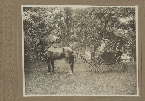 A buggy pulled by two horses stands in the woods. Two men sit in the front seat and two women in the back seat. All four are wearing hats and formal clothes.