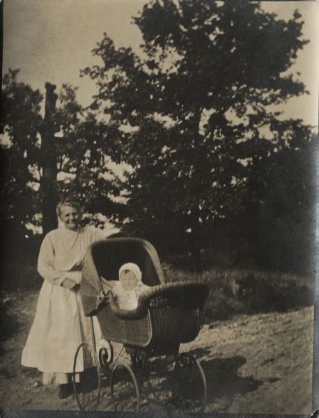 A woman pushing a young child in a baby carriage. They are outdoors with trees in the background.