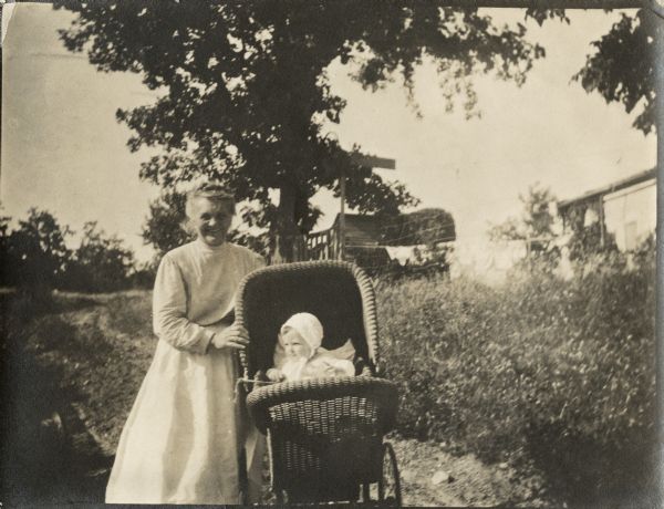 A woman pushing a young child wearing a cap in a baby carriage. There appears to be laundry hanging on a line in a yard in the background near some buildings.