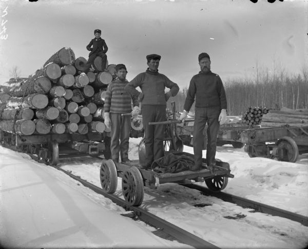 Rail siding and handcar with crew at Slashing, Waupaca County. Three men are standing on the handcar in the foreground. A young boy is sitting on a load of logs behind them.
