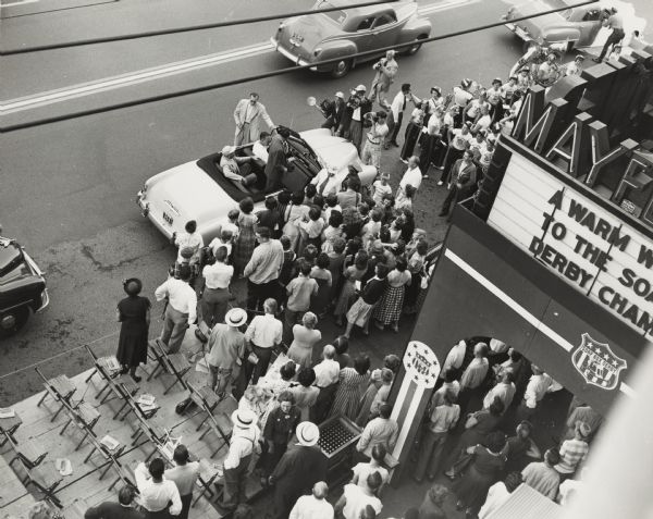 Elevated view of citizens of Akron welcoming city champions and visiting celebrities to the All-American Soap Box Derby in front of the Mayflower Hotel. A crowd is gathered in front of the hotel underneath the marquee, while photographers gather in front of an automobile where a man is standing in the back seat.