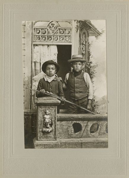 A studio portrait of two young boys, Henry Stacy (Badsoldier) and George Bearchief, standing with bow and arrows by a prop stone wall, in front of a painted backdrop. Both boys are wearing hats and handkerchiefs around their necks.