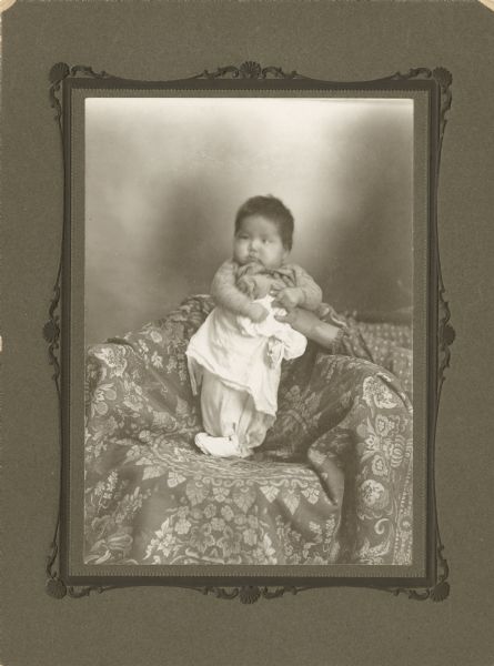 Full-length studio portrait of a Ho-Chunk baby wrapped in a white blanket. The baby is posing standing in a covered chair, and is supported by a woman beside the chair. Her arms are the only part of her body visible in the portrait.