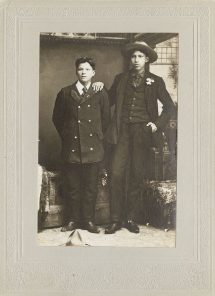 Full-length studio portrait of two young Ho-Chunk men posing standing and wearing suits and ties in front of a painted backdrop and prop stone wall. The young man on the right wearing the hat and flowers in his lapel is Henry Stacy (Bad Soldier), and the other young man is William Rainbow.