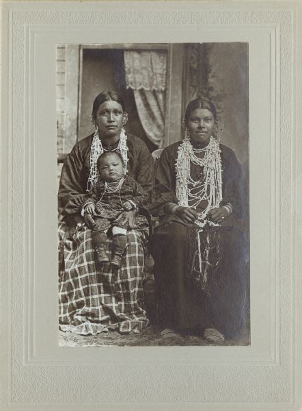Full-length studio portrait in front of a painted backdrop of two Ho-Chunk women posing sitting near a prop wood fence. They are wearing several necklaces, earrings, and file bracelets. The woman on the left is holding a child on her lap who is also wearing necklaces.