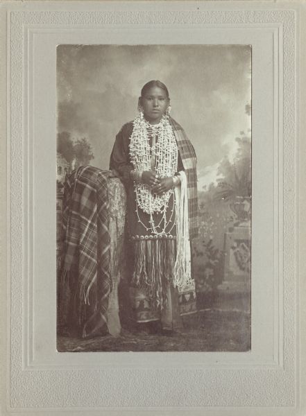 Full-length studio portrait of Henukaw Davis, Rogue. She is wearing several necklaces, earrings, rings, streamer, moccasins. A ribbon work skirt with a plaid shawl is draped on a stump. In the background is a painted backdrop.