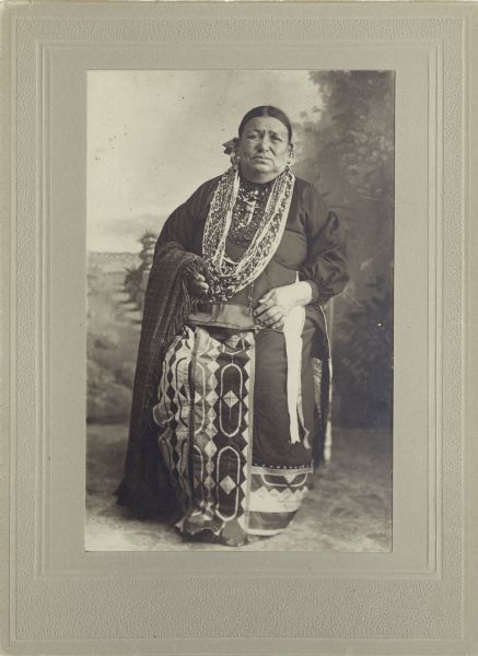 Full-length studio portrait in front of a painted backdrop of a Ho-Chunk woman, Mrs. Little Soldier, posing sitting with a plaid fringed shawl over her left arm. She is wearing several necklaces, earrings, and a ribbon work skirt. Mrs. Little Soldier was the wife of the medicine man.